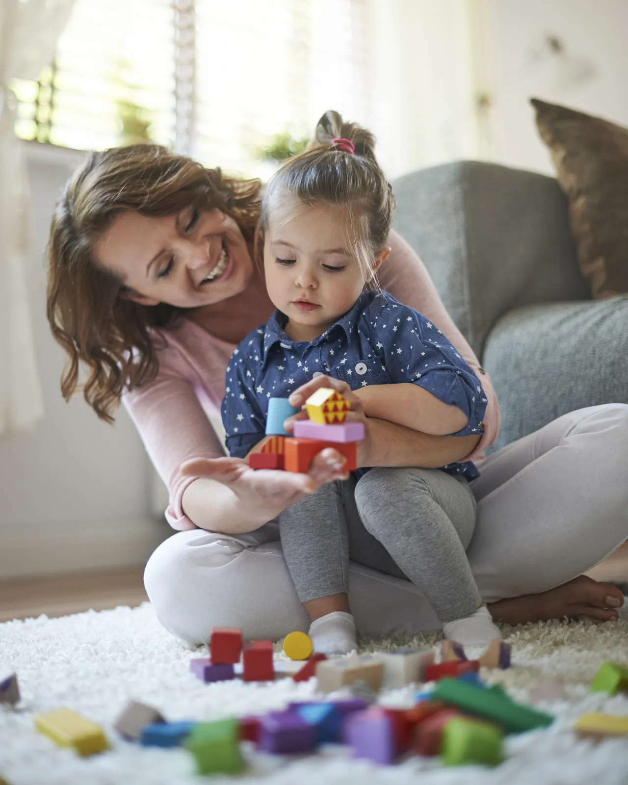A mother laughing as her young daughter builds with colorful wooden blocks